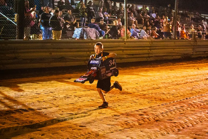 Kids racing homemade cardboard box cars down the front stretch under the lights